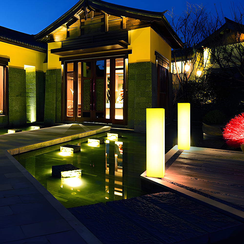 Square floor lamp casting a warm glow along a poolside pathway with modern architecture.