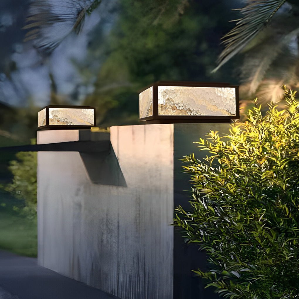 Square outdoor pillar lights mounted on a tall concrete column surrounded by greenery.
