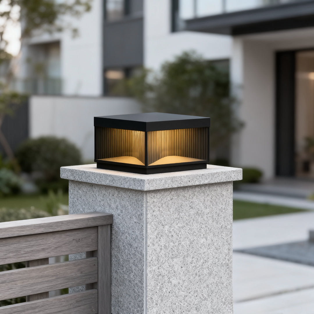 Square outdoor pillar lights installed on white stone post providing warm ambient lighting in landscaped garden daytime.