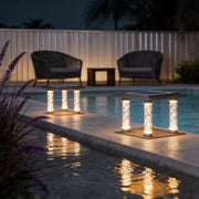 Square pillar lamp with crystal columns, providing warm illumination near a poolside seating area.