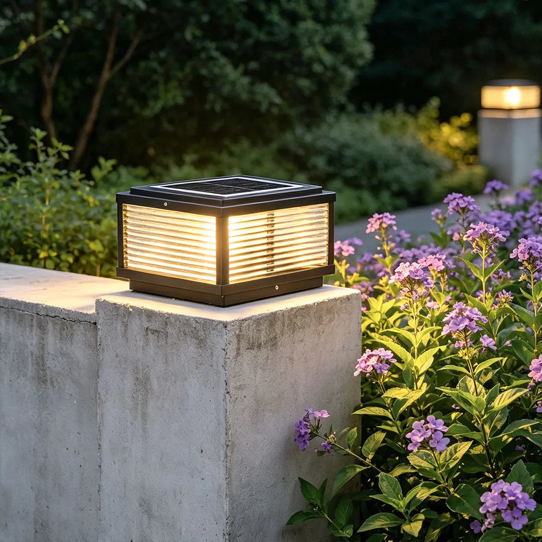 Square pillar light on a concrete fence post surrounded by flowers in a landscaped yard.
