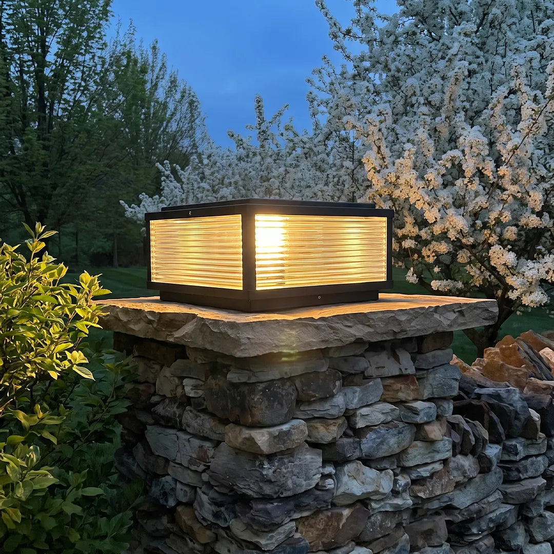 Square pillar light mounted on a stacked stone column, featuring a black frame, ribbed diffuser panels, and warm outdoor illumination.