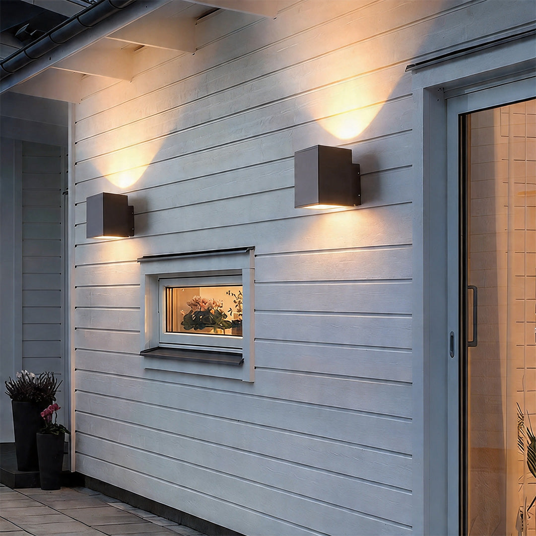 Square wall sconce up and down light fixtures glowing on a white horizontal siding house wall.