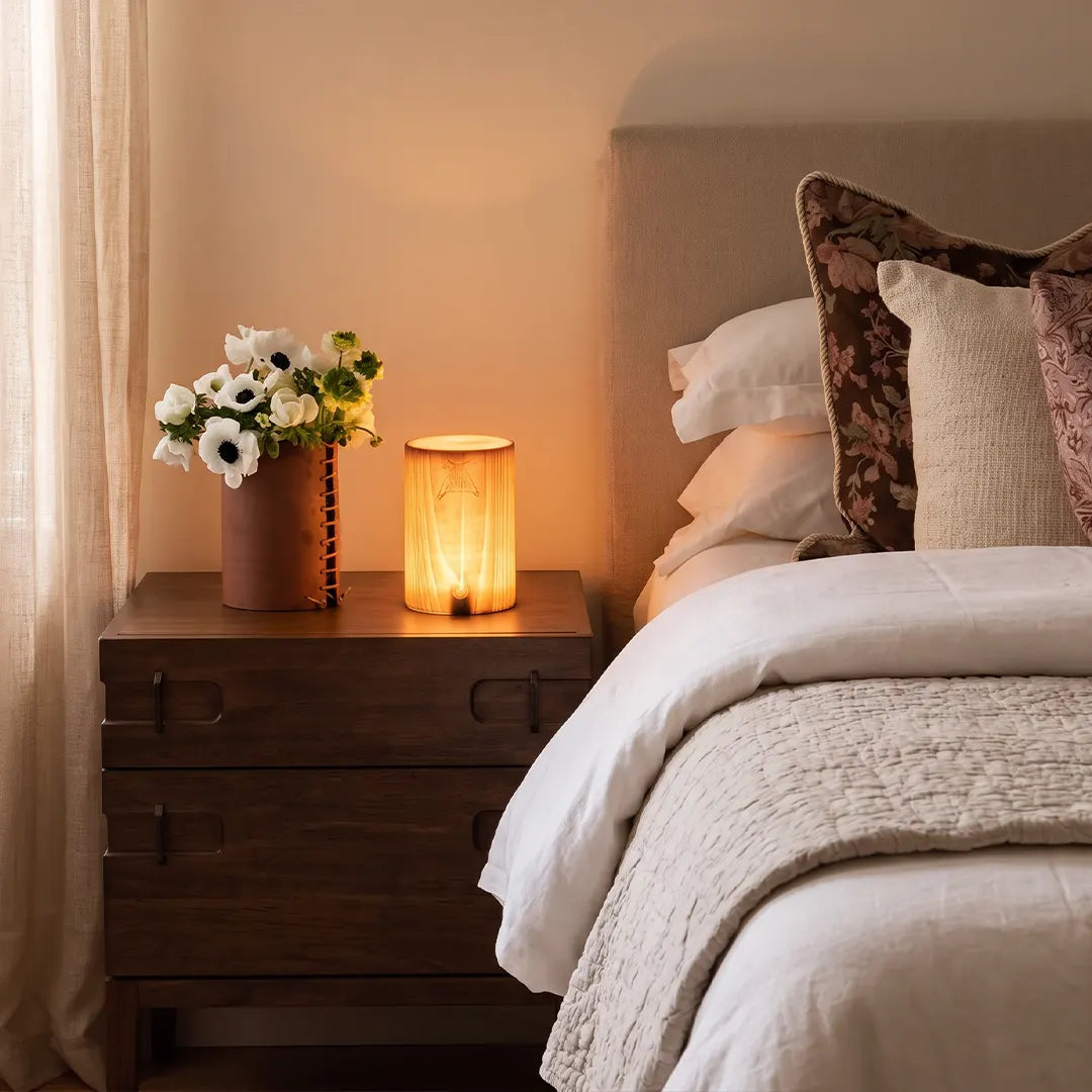 Square wood lamp with a warm glowing shade styled on a dark wood nightstand beside a cozy bed.