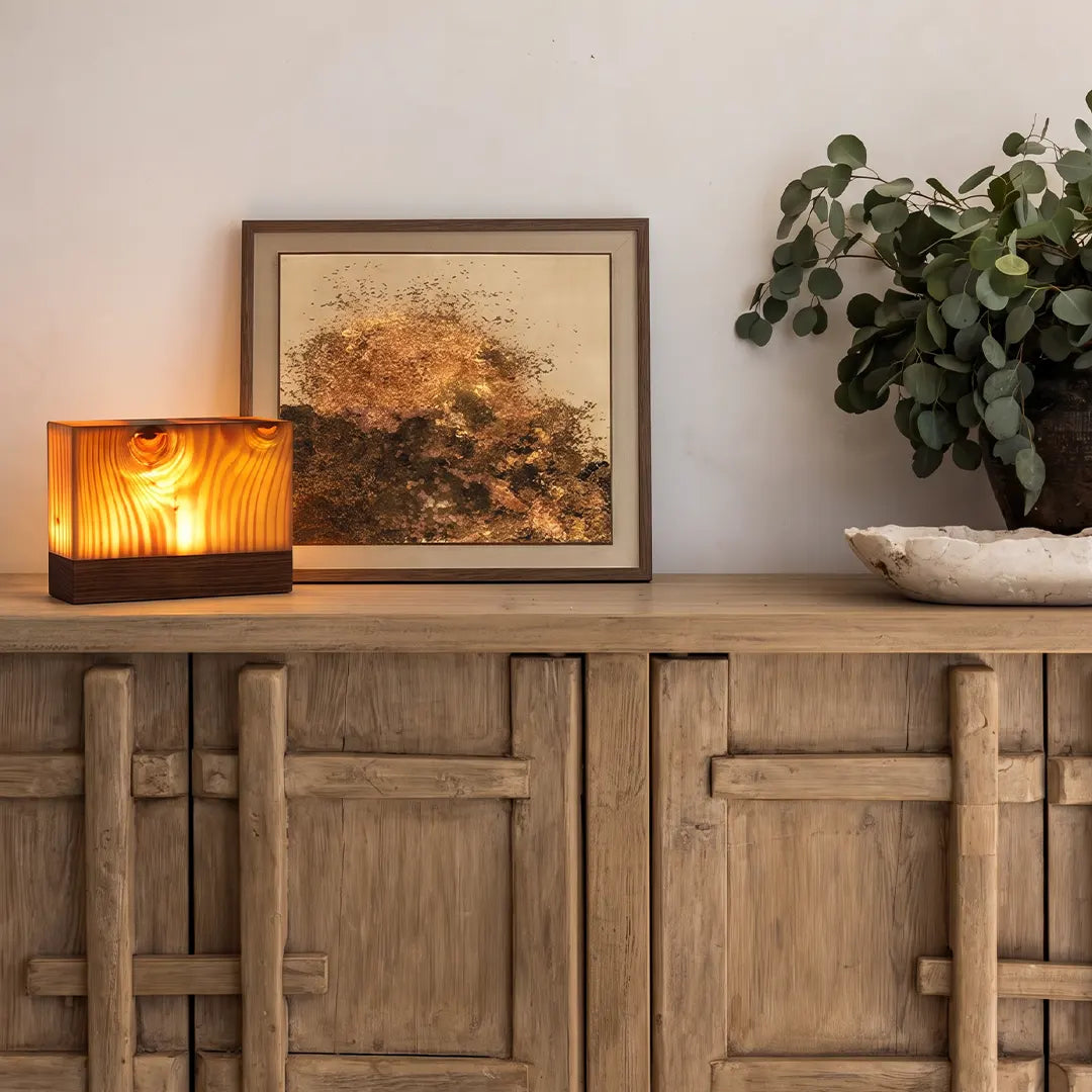 Square wood lamp with a warm ambient glow displayed on a rustic wood sideboard with framed artwork and greenery.