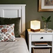 Square wood lamp with a warm glow, placed on a wooden nightstand in a cozy bedroom.