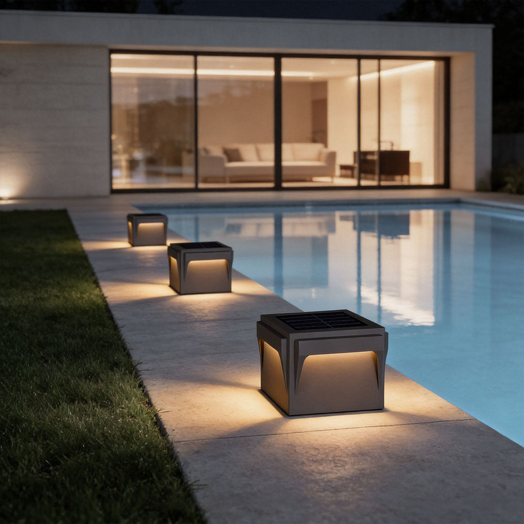 Modern stainless steel outdoor pillar lights illuminating a pathway beside a luxurious pool and contemporary house.