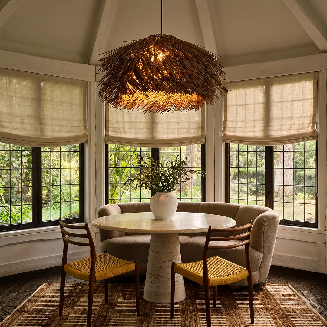 Straw pendant light hanging above a round dining table in a bright breakfast nook with large windows.