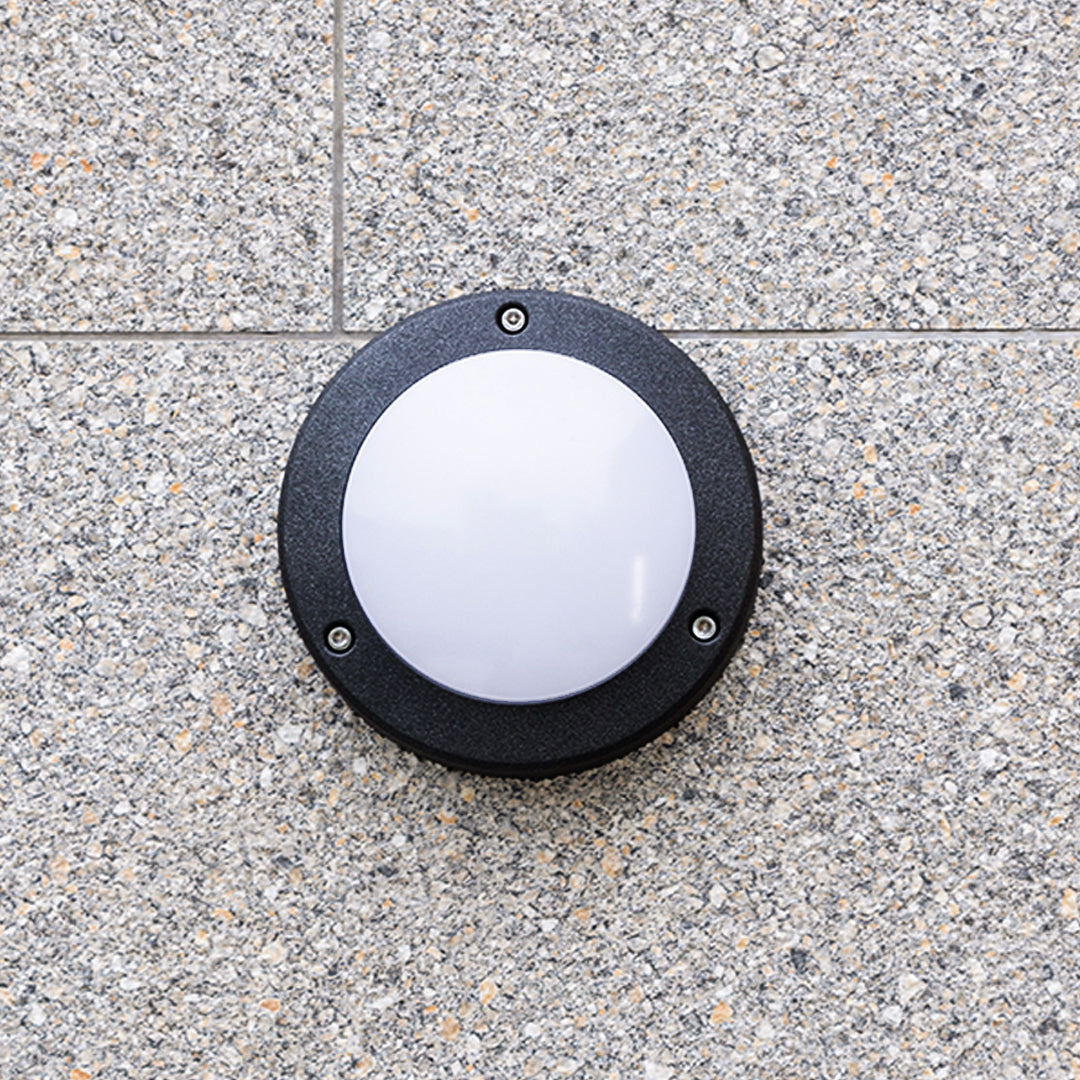 Close-up of a sturdy black round outdoor wall lights fixture with a white frosted diffuser mounted on textured aggregate wall.
