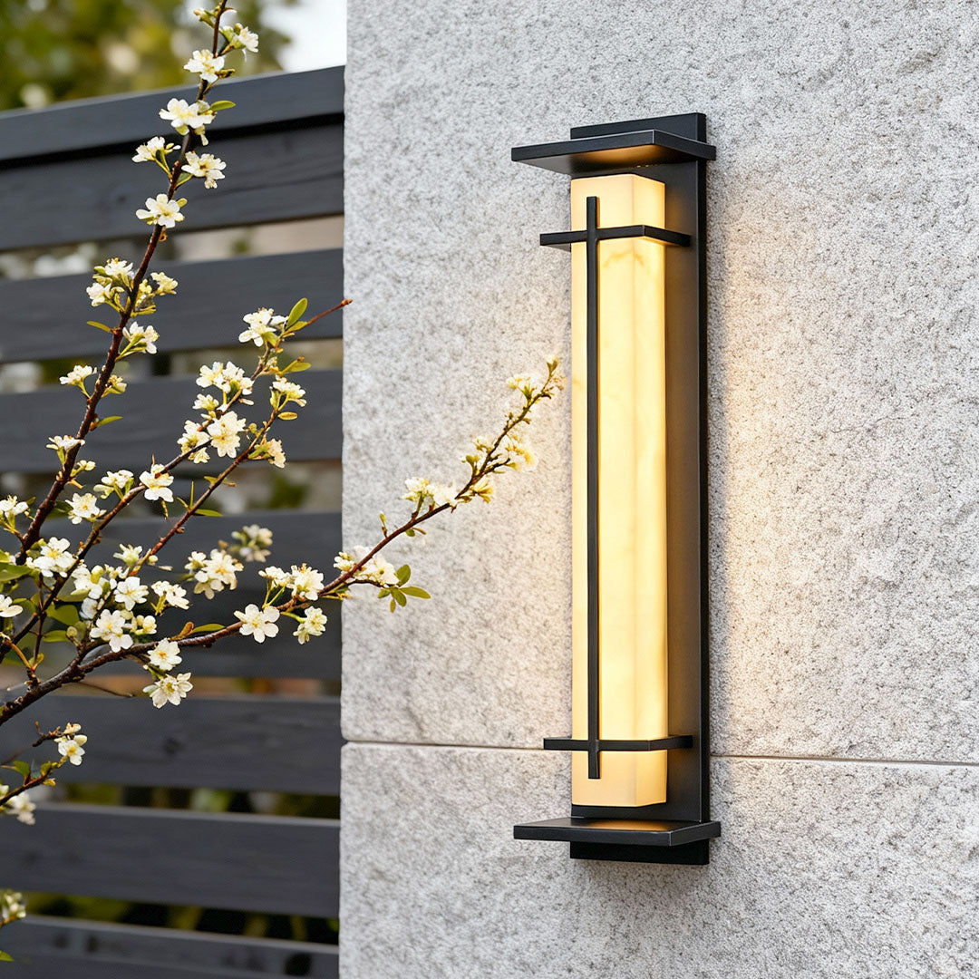 Close-up of a stylish black outside wall light with a frosted diffuser on a granite wall next to blooming branches.