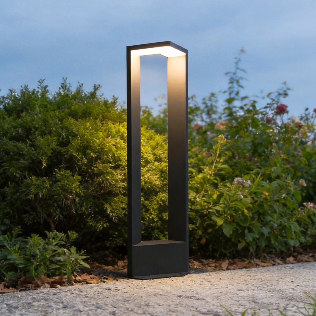 Tall modern architectural garden path light glowing warmly next to a gravel walkway and bushes.