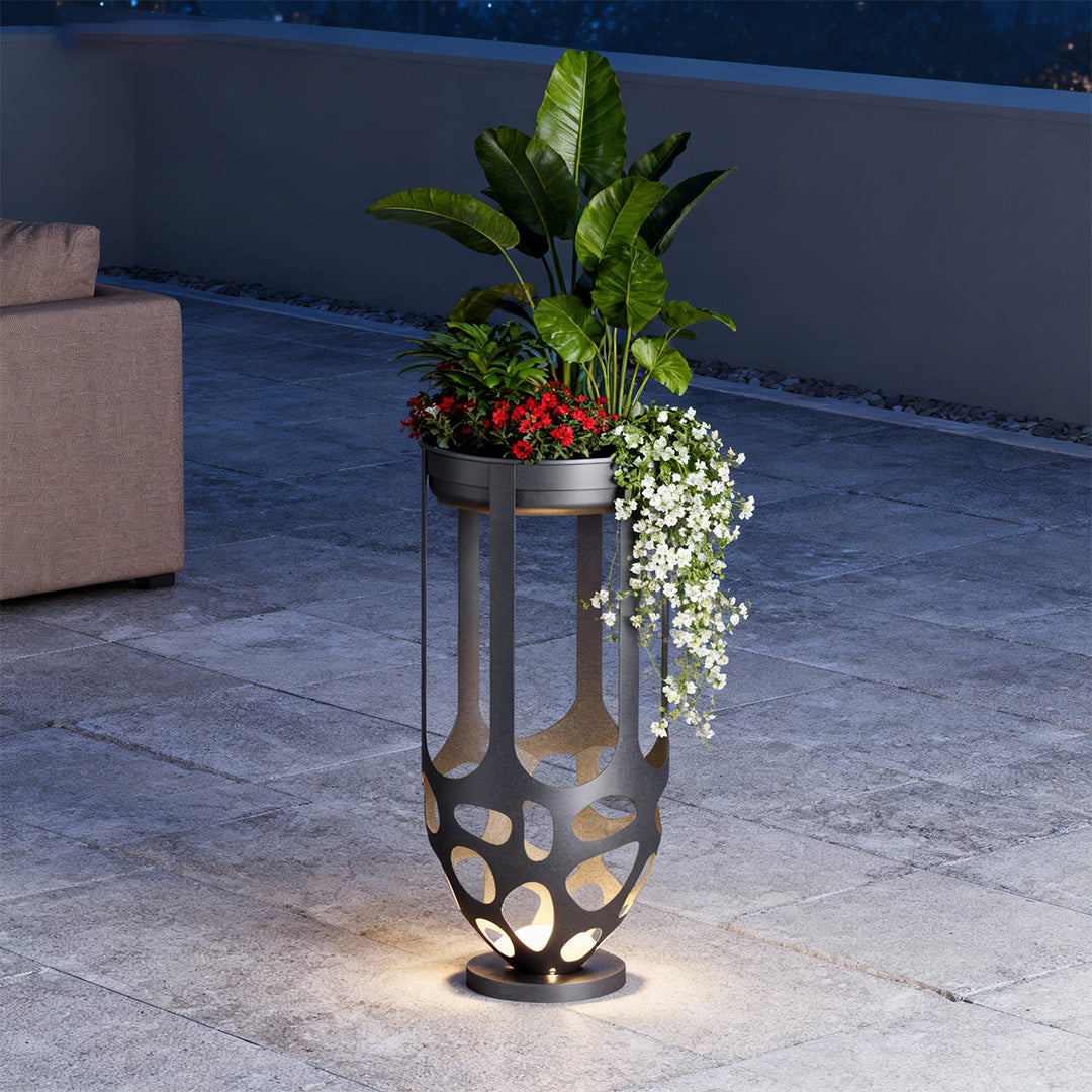 Tall black metal lighted outdoor planter displaying vibrant flowers on a patio at dusk.