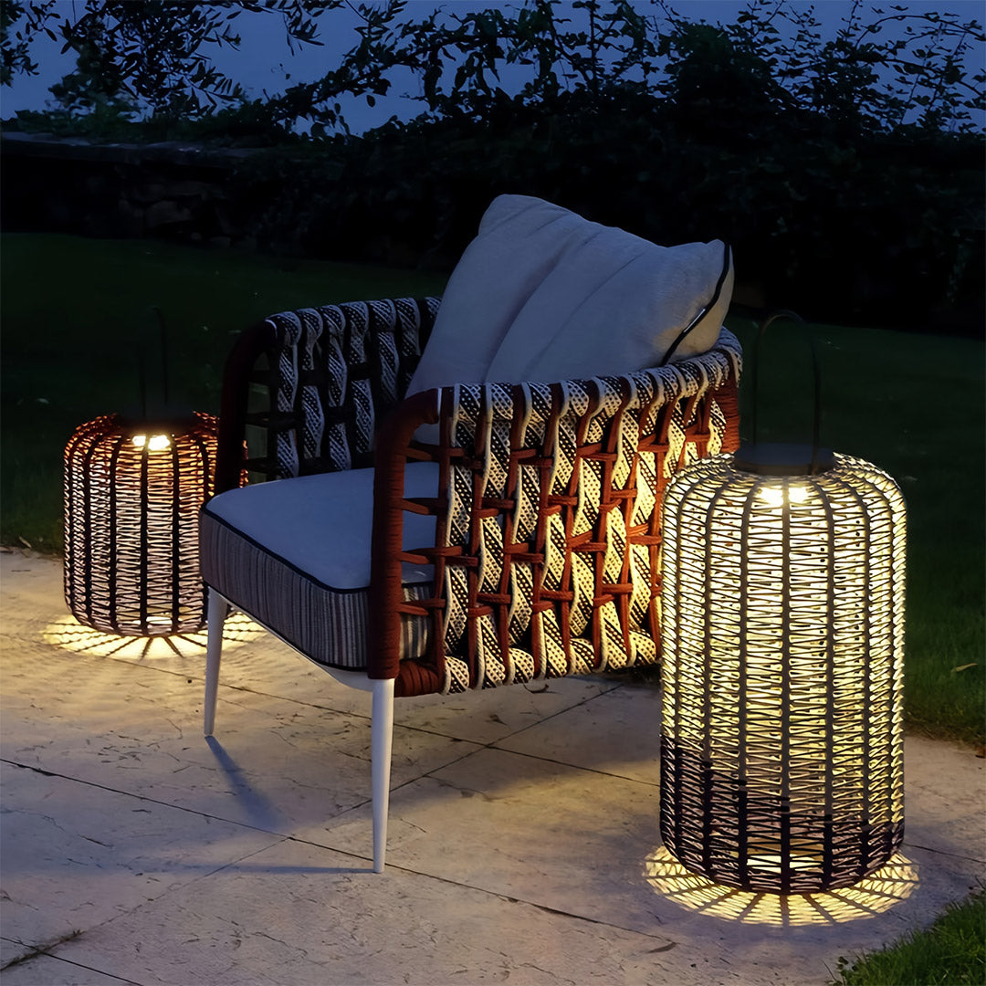 Outdoor LED lantern with a tall, cylindrical woven resin design, providing ambient light next to a woven armchair on a patio.