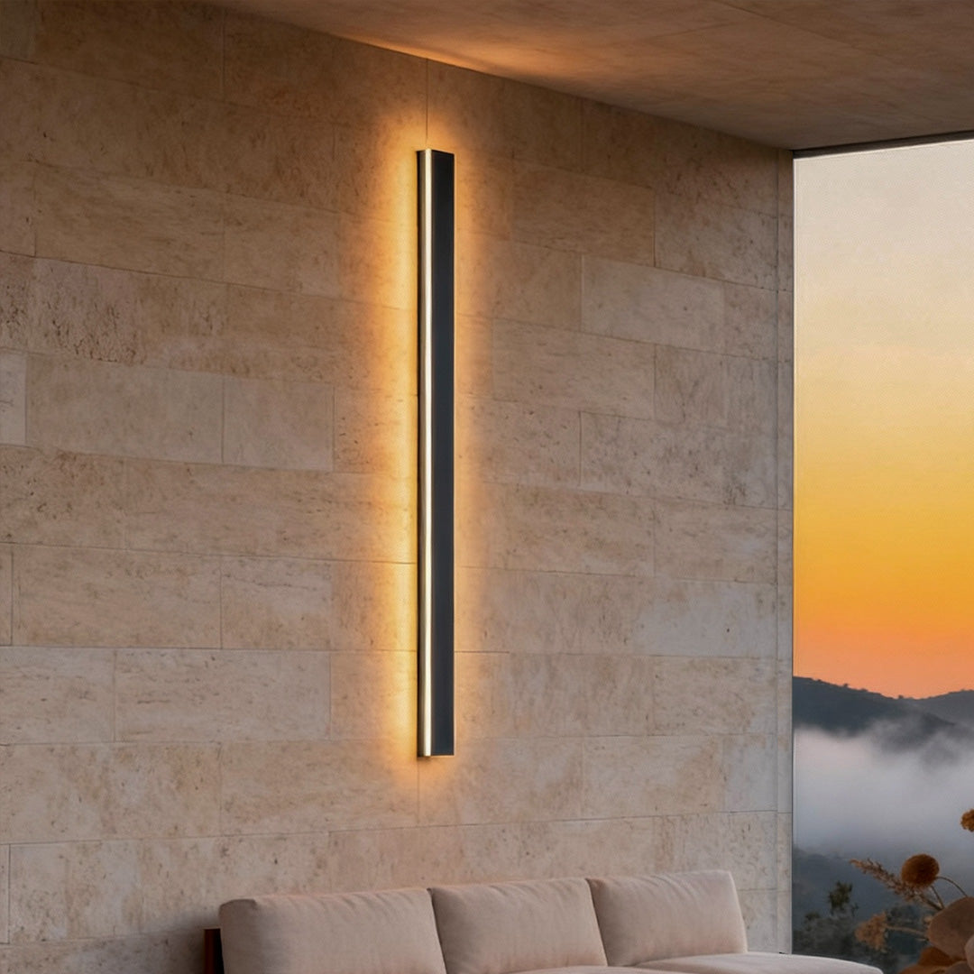 Tall linear outdoor wall sconce with warm LED light, mounted on textured stone wall for contemporary exterior lighting.