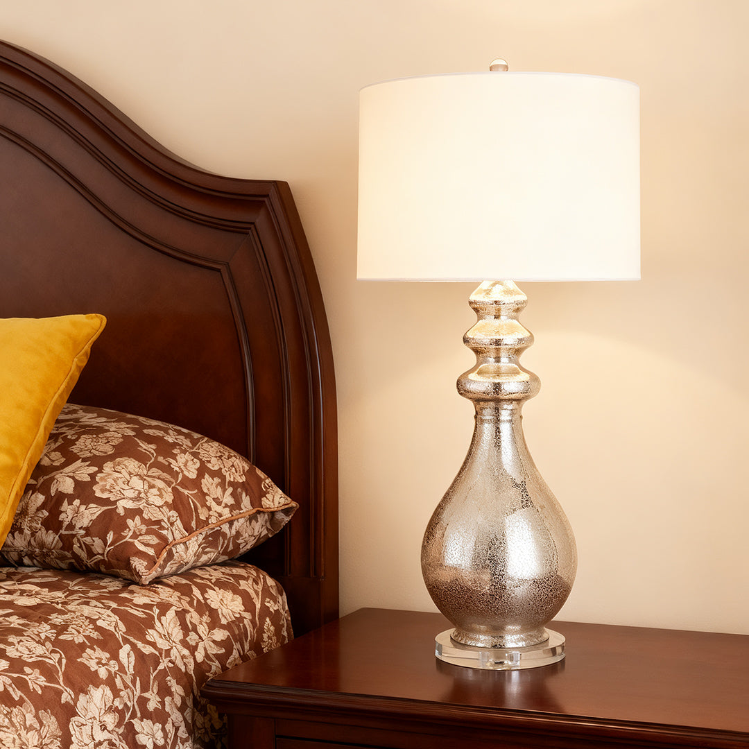 Tall, mercury glass modern silver table lamp illuminating a rich, dark wood nightstand in a classic bedroom setting.