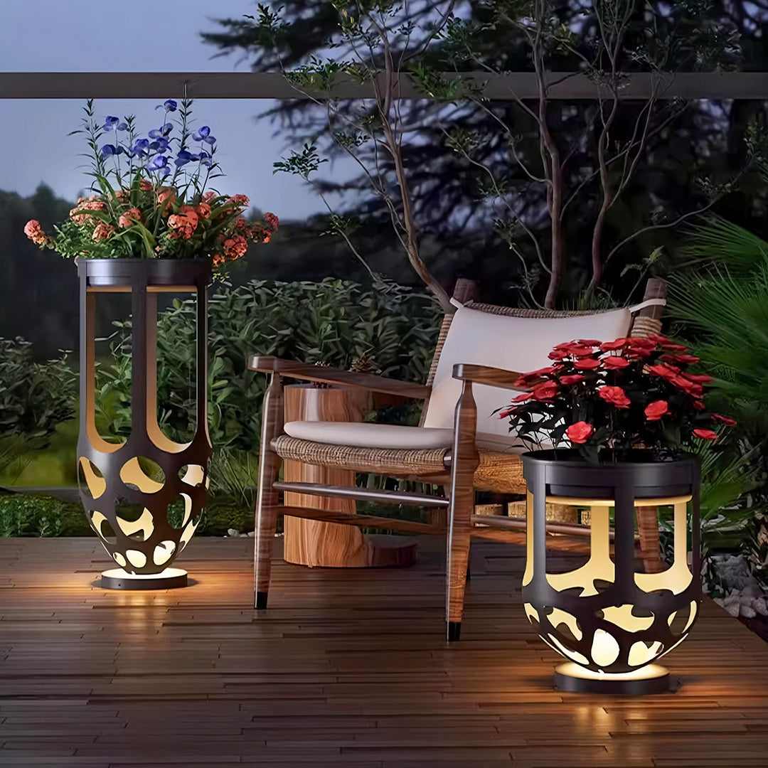 A set of tall and short lighted outdoor planter stands holding flowers and illuminating a wood patio at dusk.