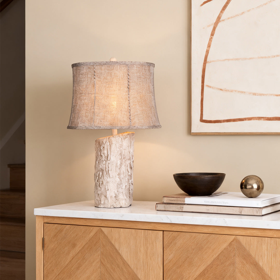Tall white wood table lamp on a light-colored wooden sideboard, featuring a simple silhouette and neutral textured drum shade.