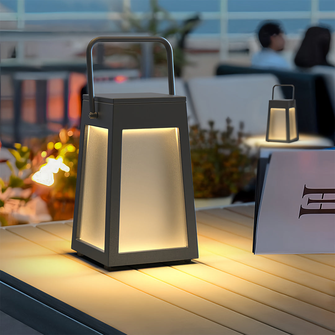Tapered square camping lantern placed on a wooden table at an outdoor cafe or restaurant patio, providing warm table light.