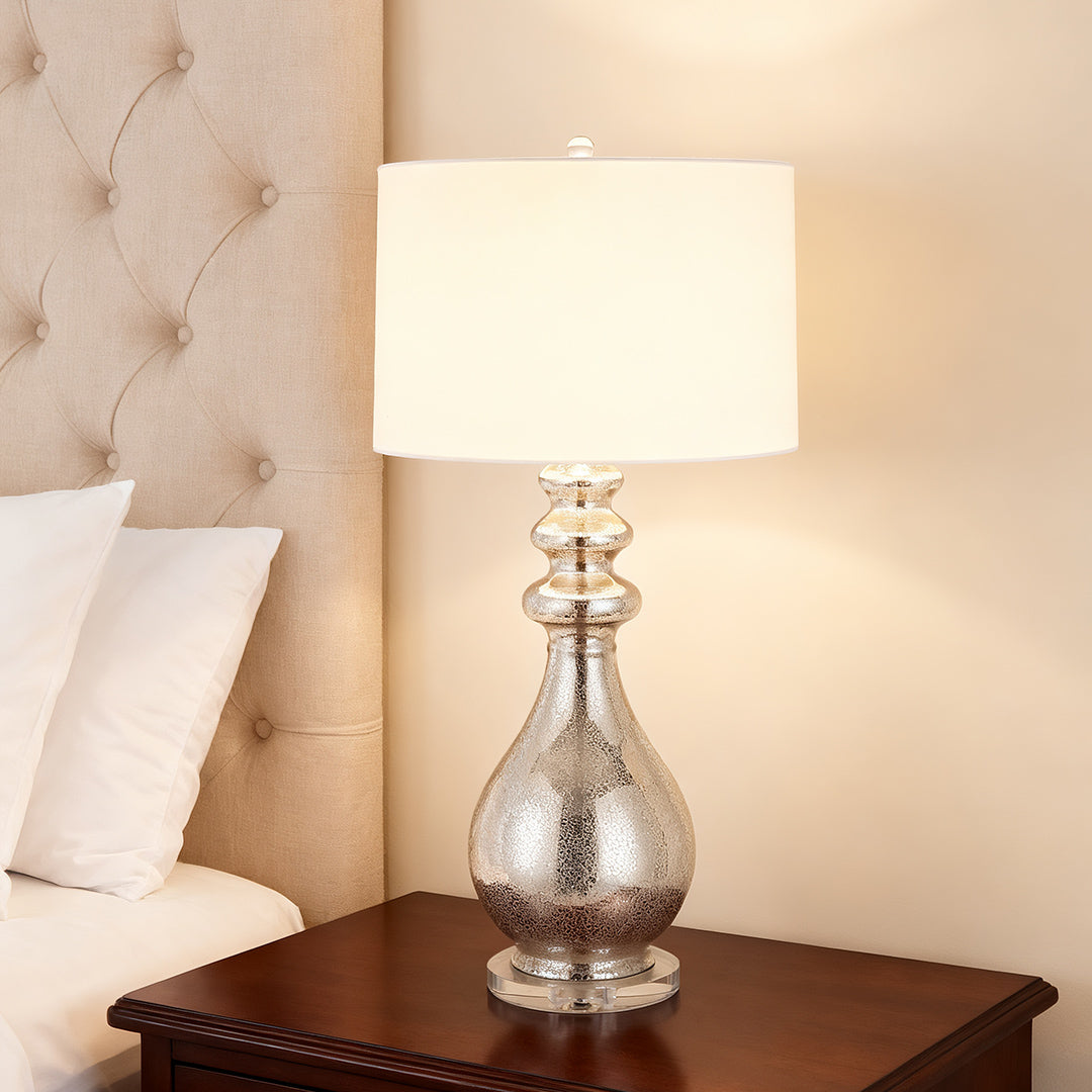 Textured glass base modern silver table lamp with a white drum shade, providing soft light next to a tufted headboard.