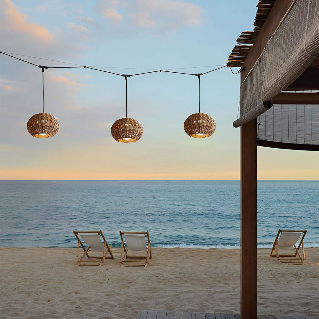 Three rattan outdoor pendant lights hanging on a scenic beach with lounge chairs during sunset.