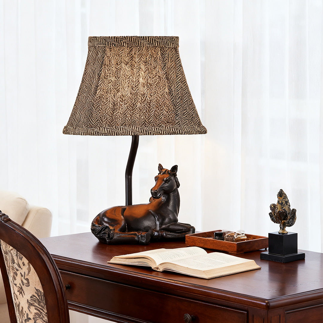 Traditional table lamp horse sculpture base with a patterned fabric shade, placed on a dark wood desk next to an open book.