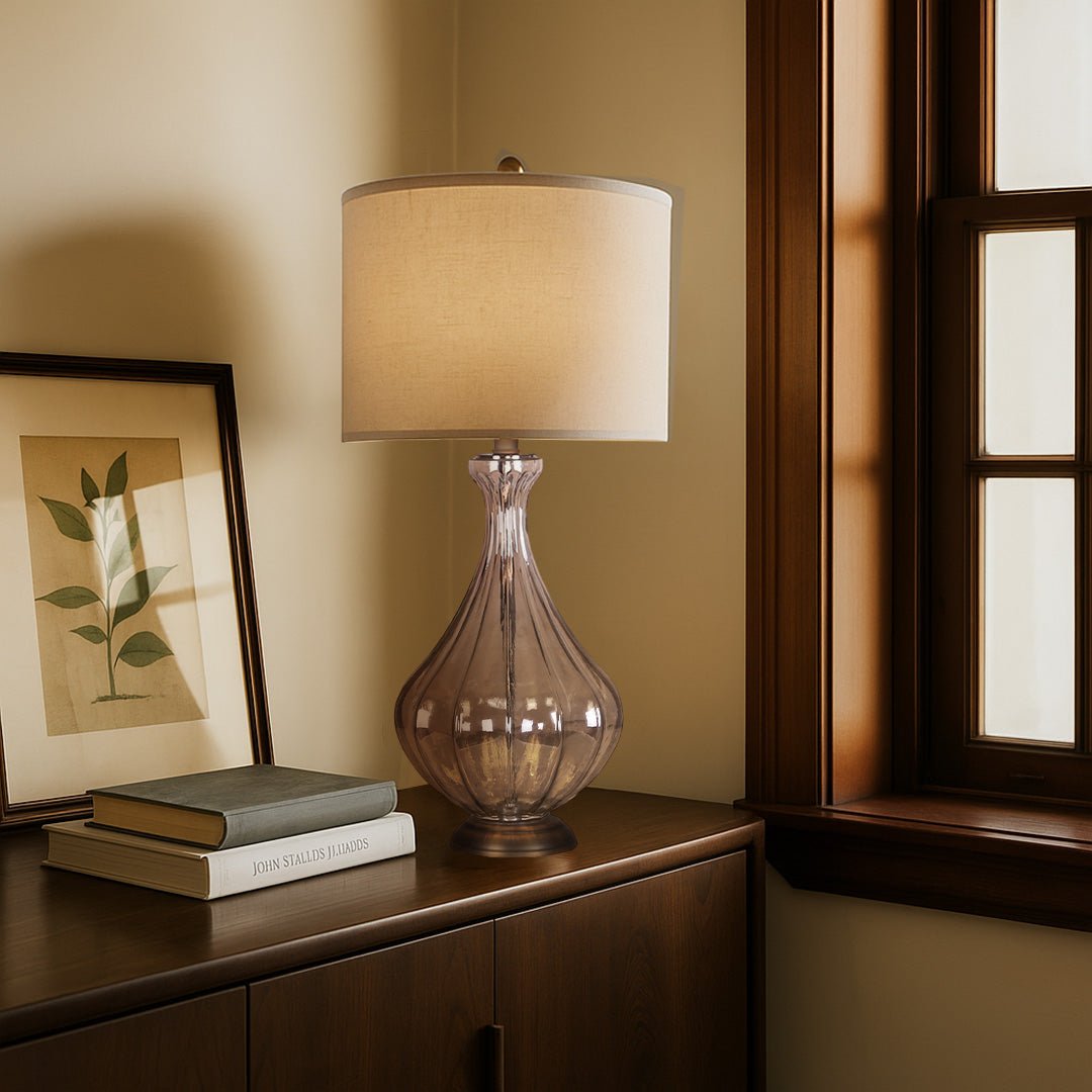 Transitional glass base table lamp providing a soft, warm glow on a dark wood dresser with framed artwork.