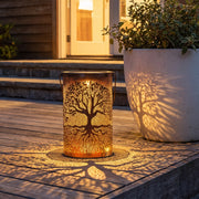 Tree of life lantern glowing on a wooden patio and casting branch-shaped shadows beside a planter.