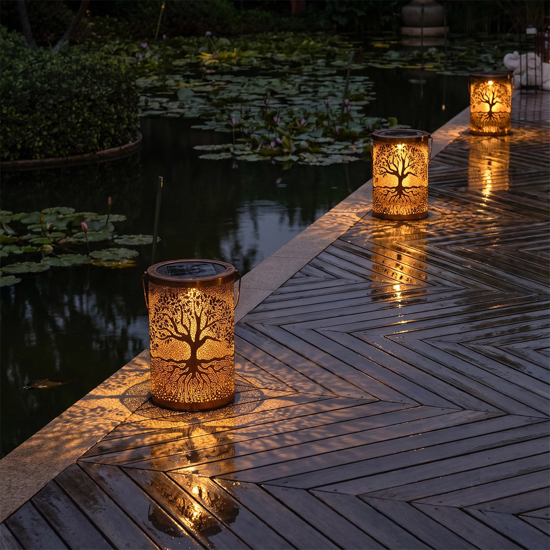 Tree of life lanterns lining a wooden deck beside a pond, casting warm decorative patterns.