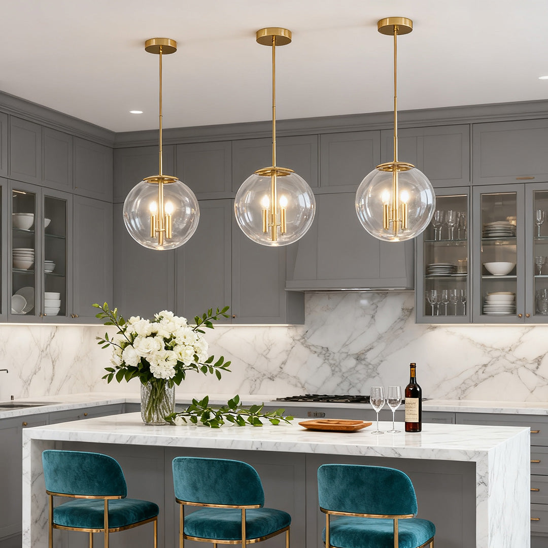 Trio of contemporary clear globe pendant light fixtures with brass accents hanging over a gray kitchen island with white marble top.