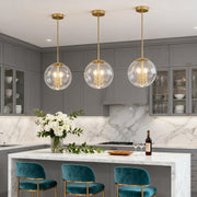 Trio of contemporary clear globe pendant light fixtures with brass accents hanging over a gray kitchen island with white marble top.