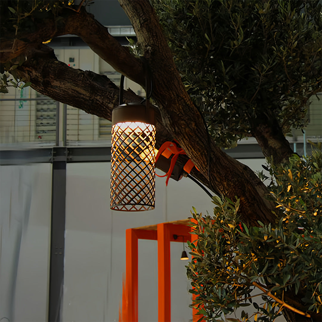 Two modern black outdoor pendant lantern fixtures suspended from a large tree, providing ambient light for an evening gathering.