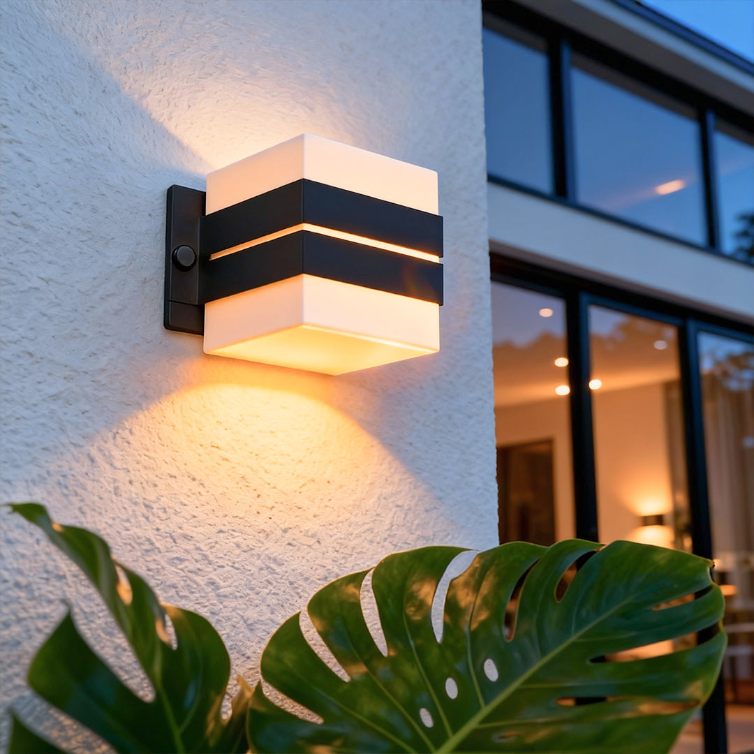 Contemporary outdoor wall sconce lighting casting up-and-down glow on house exterior beside greenery.