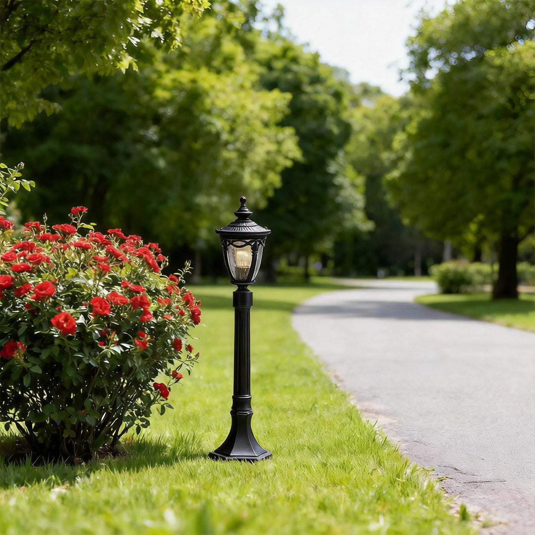 A vintage style outdoor light post standing beside an asphalt path and red rose bushes in park