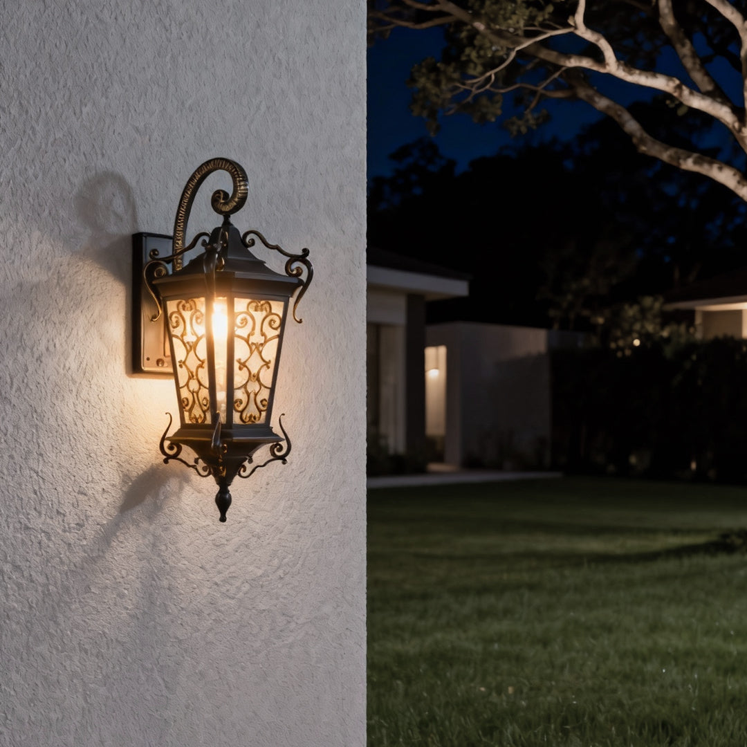 Vintage outdoor wall lantern with decorative scrollwork casting warm light on exterior stucco wall at night.