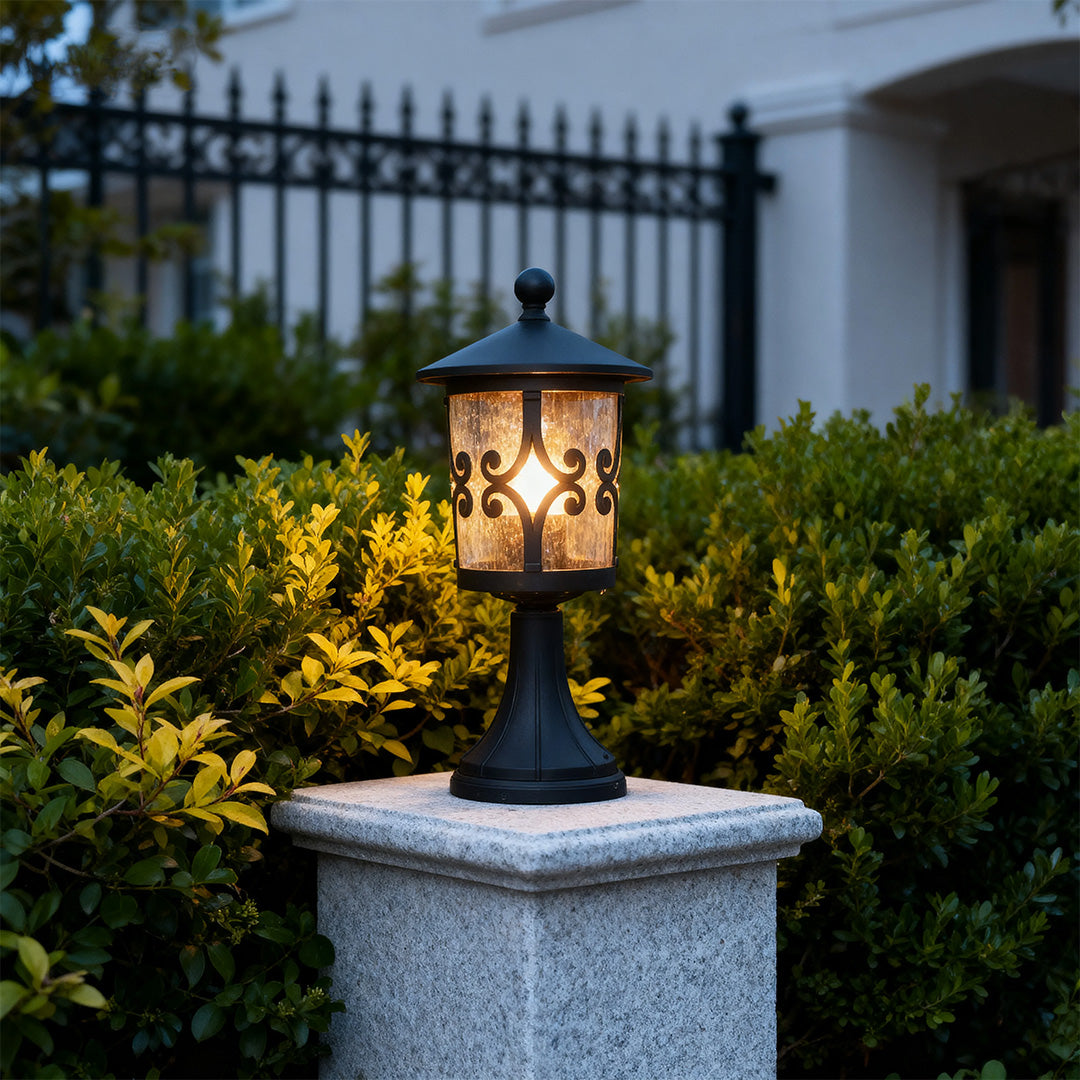 Vintage style porch pillar light glowing on granite pedestal with black iron railing and garden backdrop