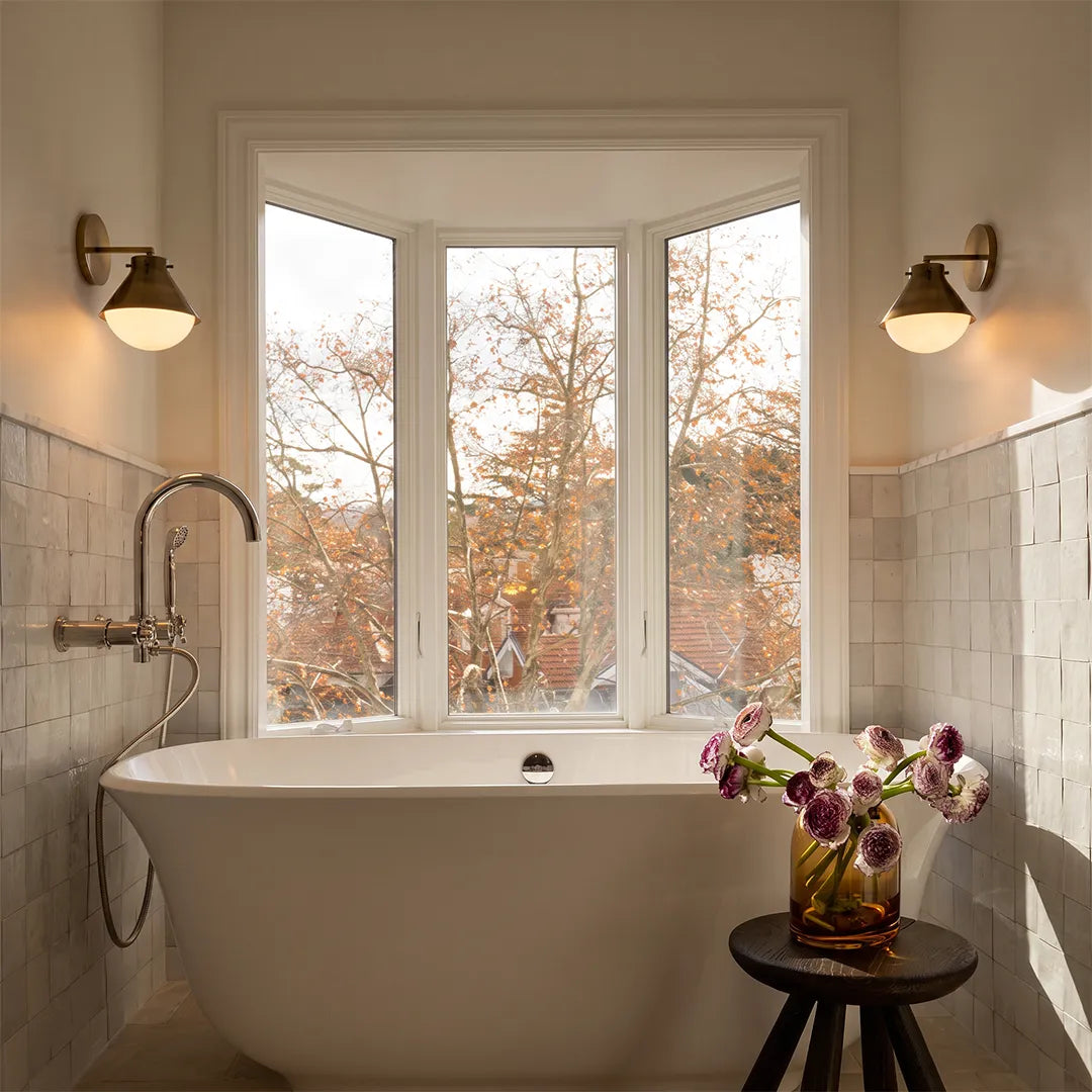 Vintage wall sconce lights mounted beside a bathtub window in a bright bathroom with warm ambient glow.