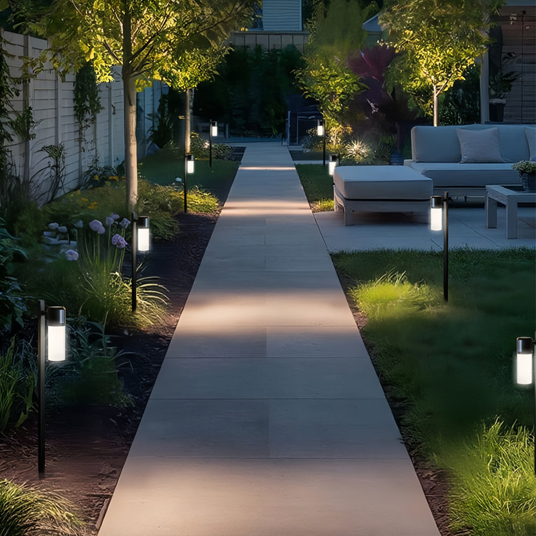 A row of contemporary path light fixtures illuminating a modern walkway lined with trees at dusk.