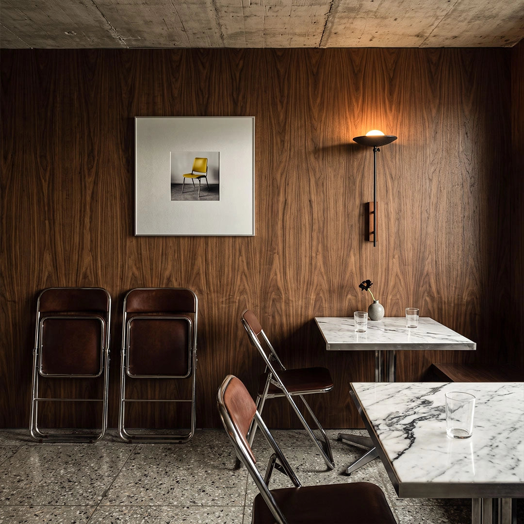 Wall adjustable light illuminating a dark wood dining nook with marble tables and black chairs.