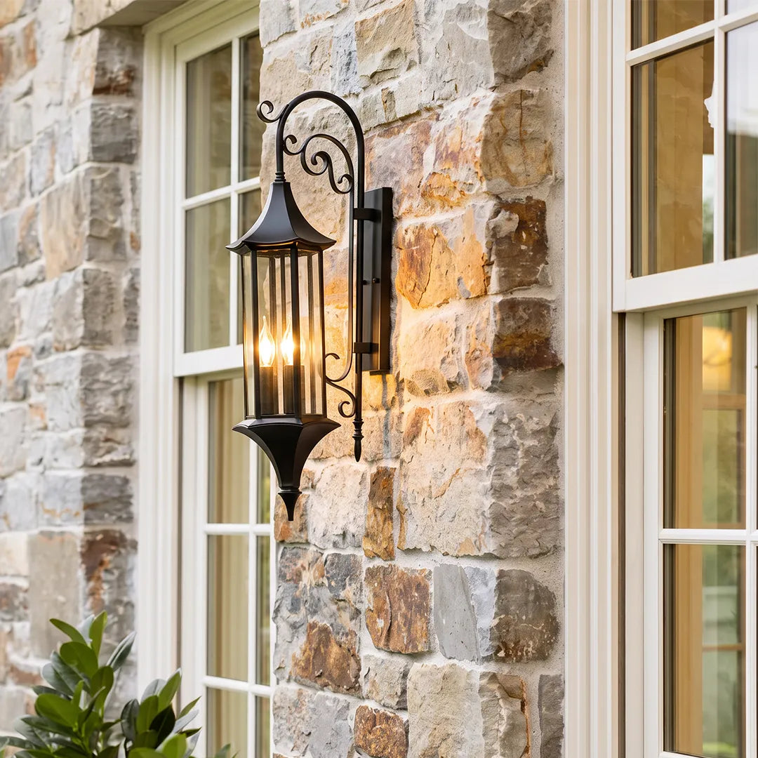 Wall lanterns outdoor lighting mounted on a stone exterior wall beside white-trimmed windows in a warm bronze finish.