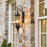 Wall lanterns outdoor lighting mounted on a stone exterior wall beside white-trimmed windows in a warm bronze finish.