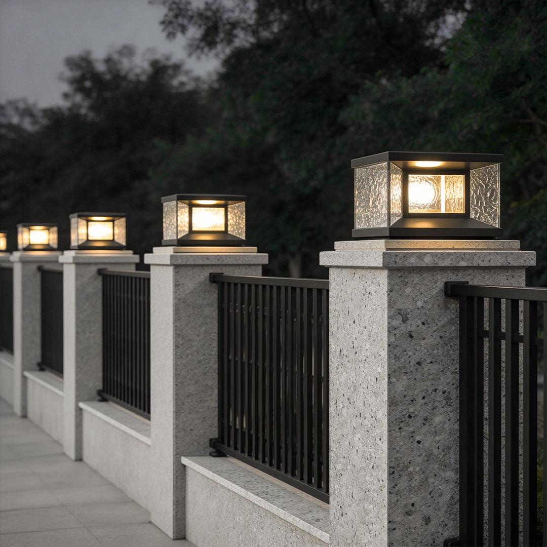 Wall pillar lights illuminating a driveway with stone pillars and black metal fence.