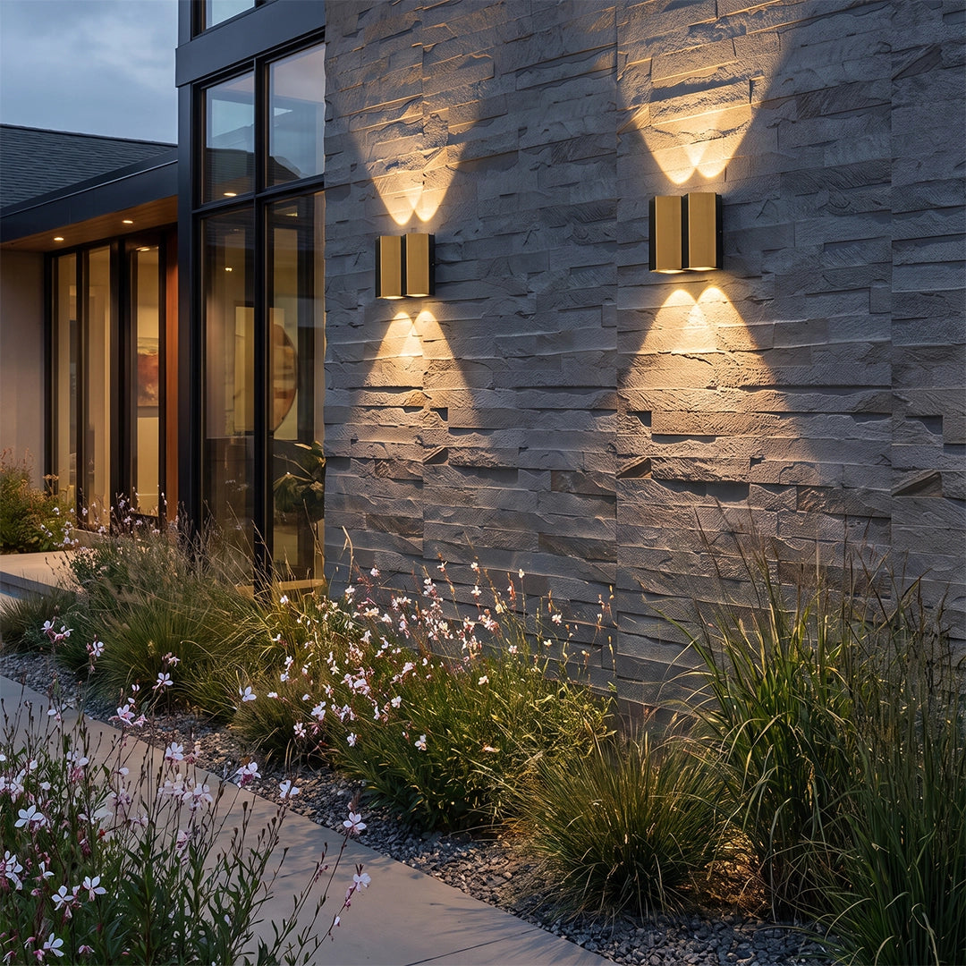 Wall sconce up-down lights installed on a stone exterior wall beside a modern garden path.