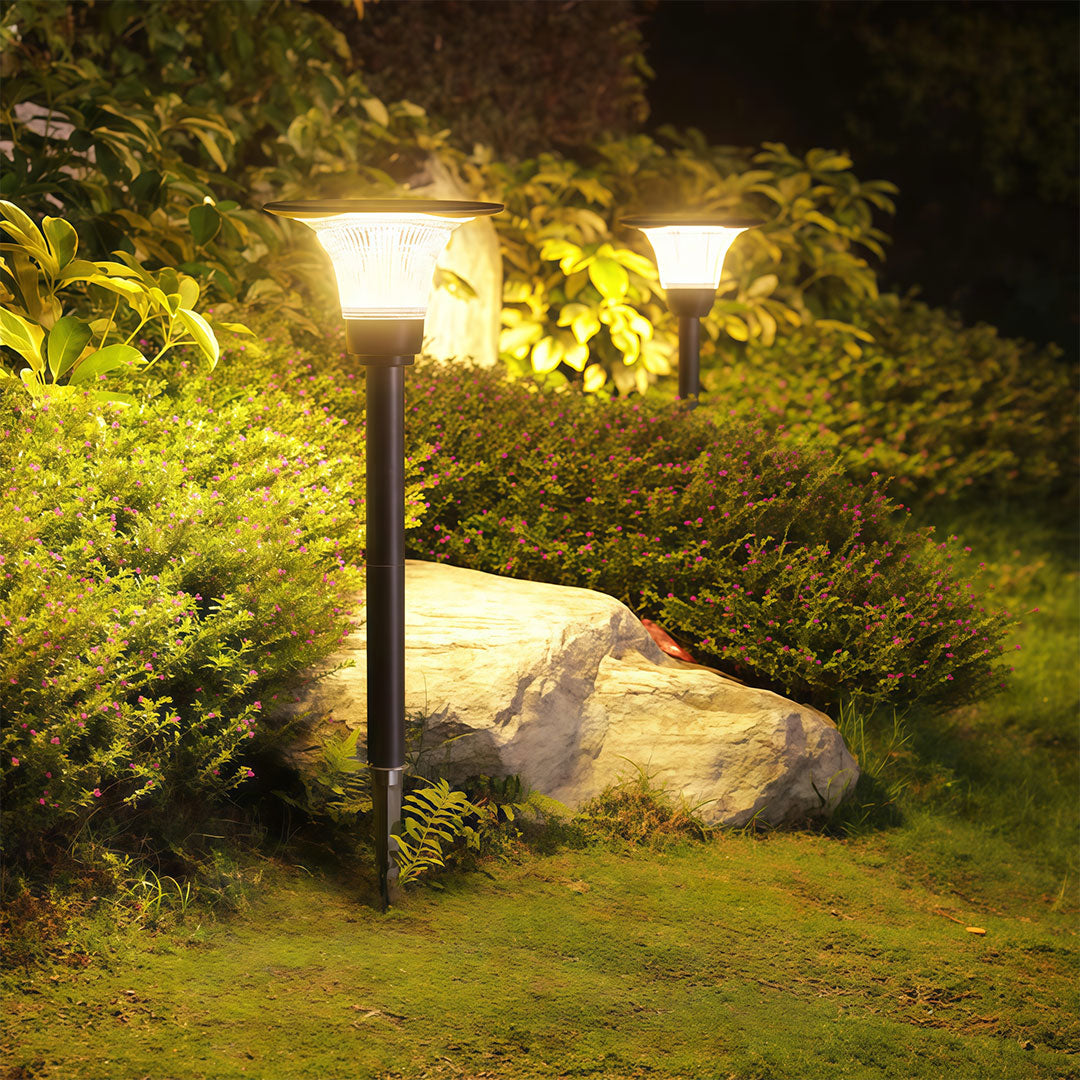 Warm-glowing black solar pathway lights illuminating a lush garden bed with rocks and shrubs at night.