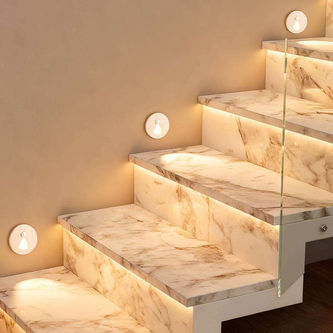 Warm interior stair step lights installed in the riser panel of a marble staircase with a glass railing, providing essential safety illumination and a luxurious accent.