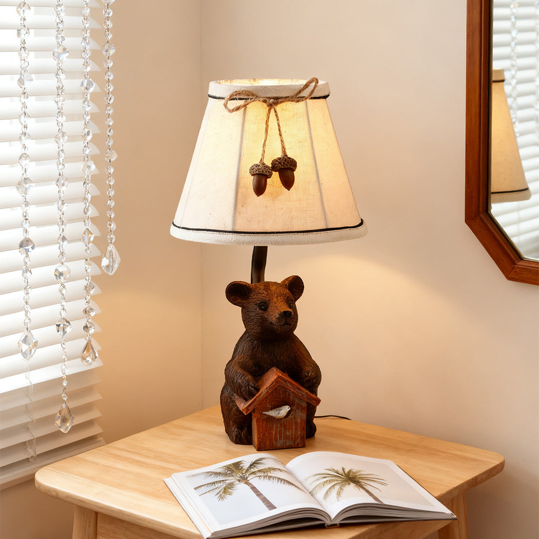 The bear table lamp casting warm light on a white-shaded desk in a cozy bedroom setting, complete with a rope-trimmed lampshade.