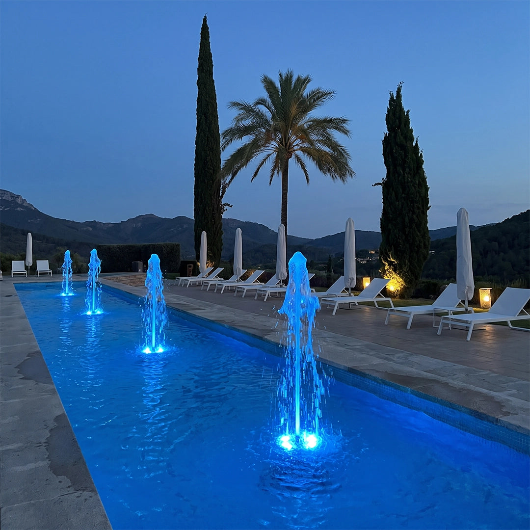 Water fountain lights producing bright blue illuminated water jets in a modern pool surrounded by palm trees.