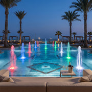 Water fountain lights illuminating multiple colorful jets across a resort pool at dusk.
