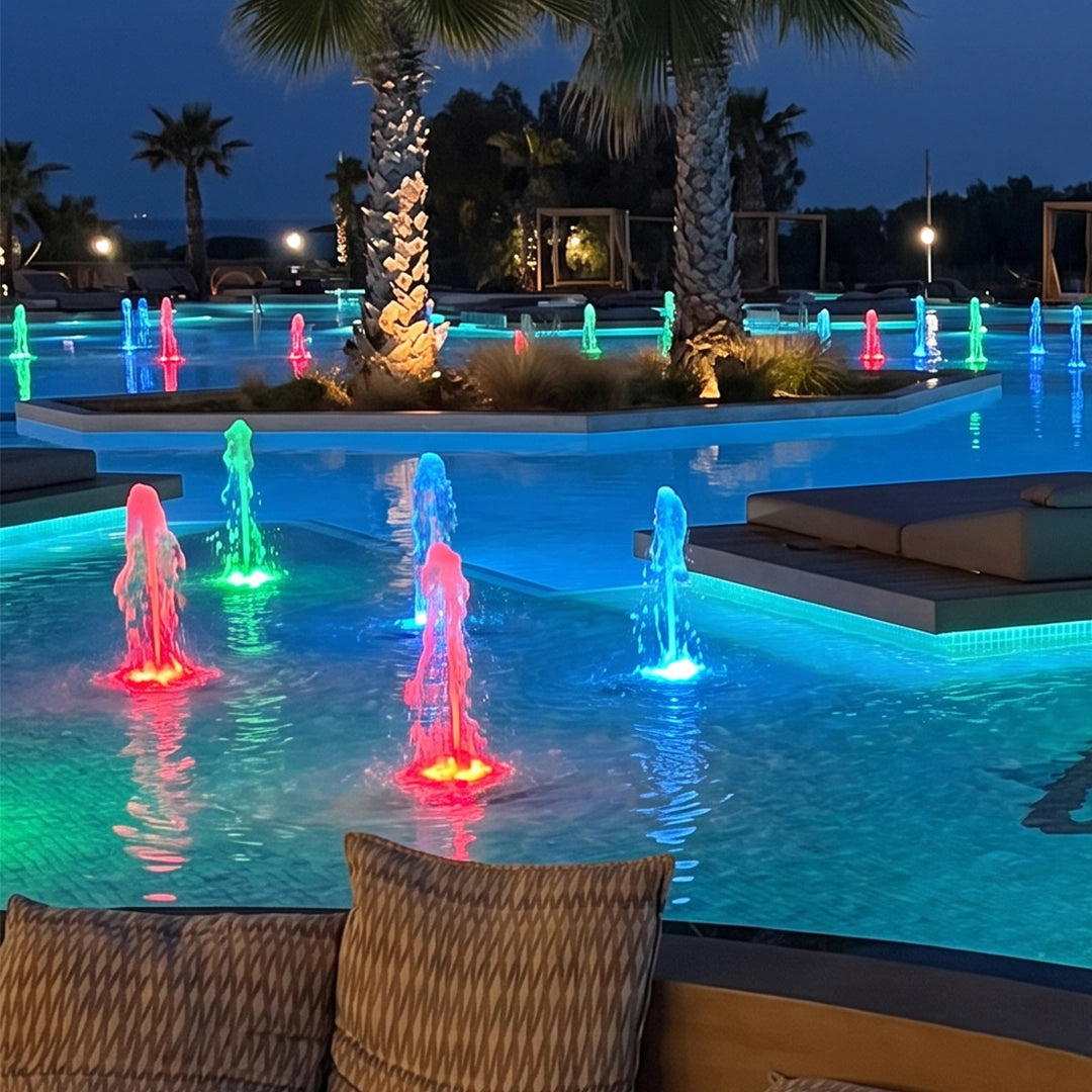 Water fountain lights adding colorful illuminated jets around a landscaped pool with palm trees at night.