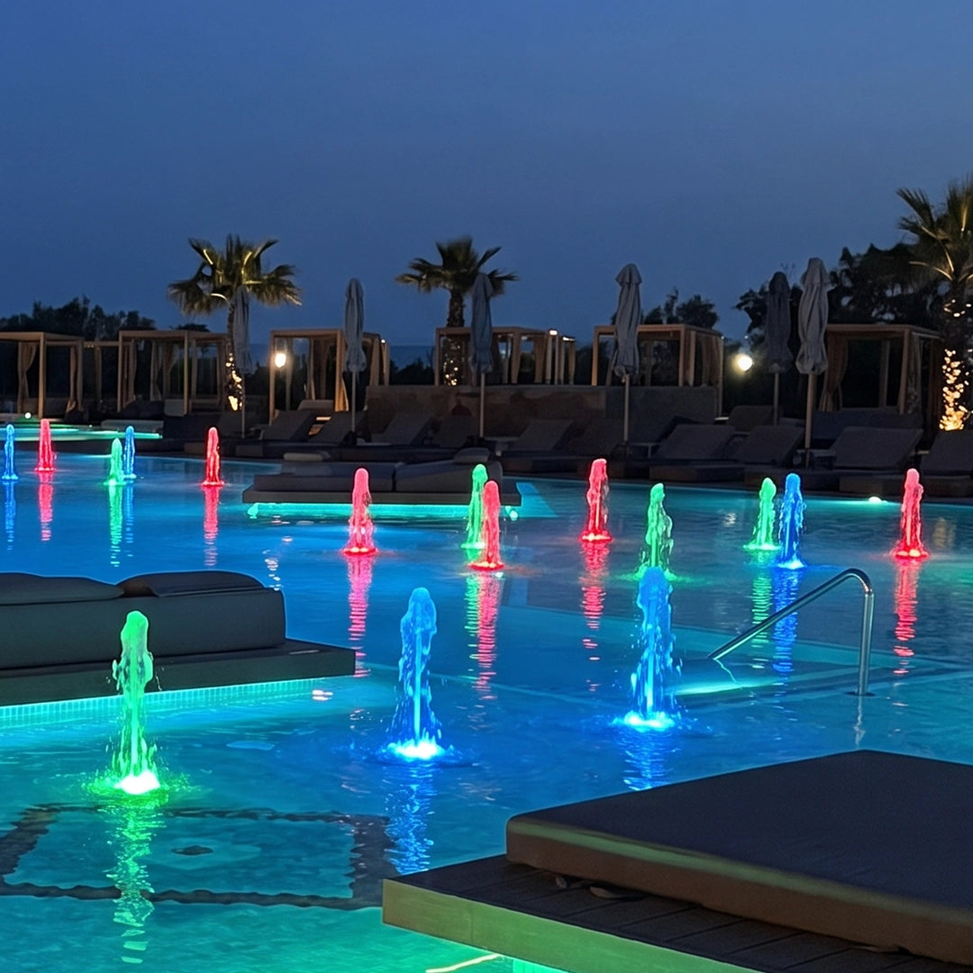 Water fountain lights creating red, green, and blue illuminated jets across a large outdoor pool.
