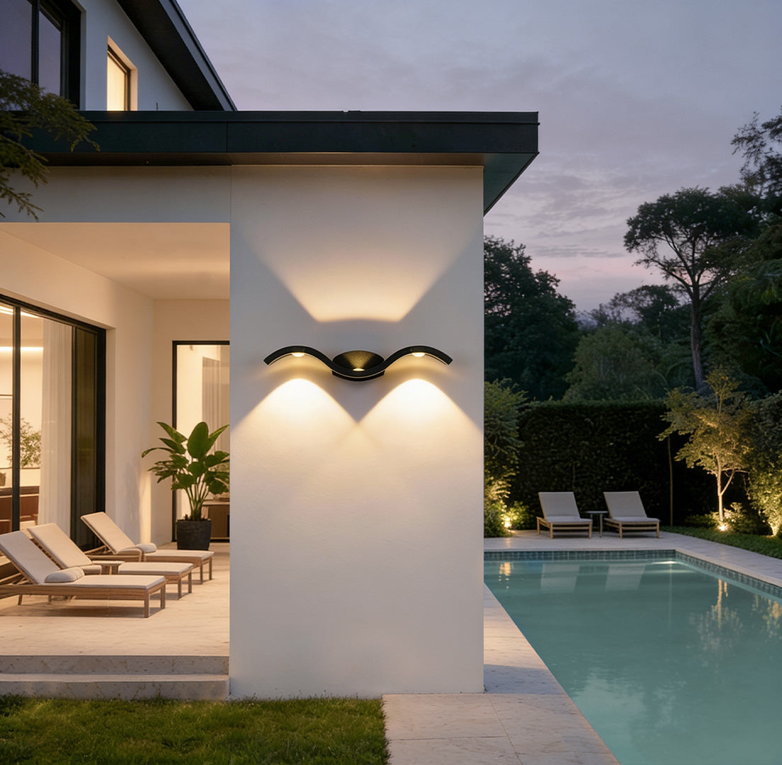 Wave wall sconce installed on a modern outdoor patio wall, casting soft up and down lighting near a pool area at dusk.